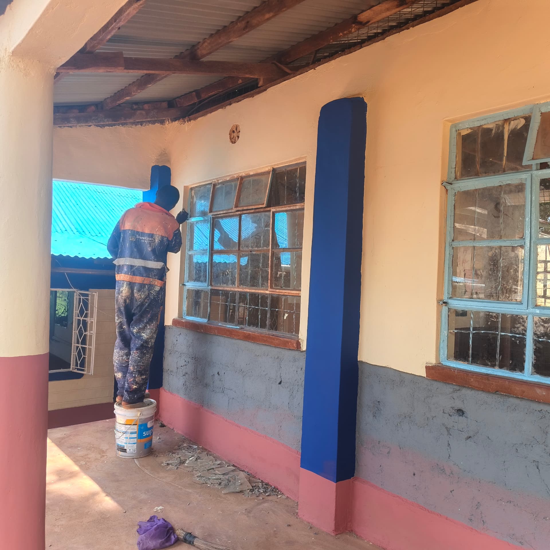 Worker painting a veranda pillar deep blue along the classroom corridor