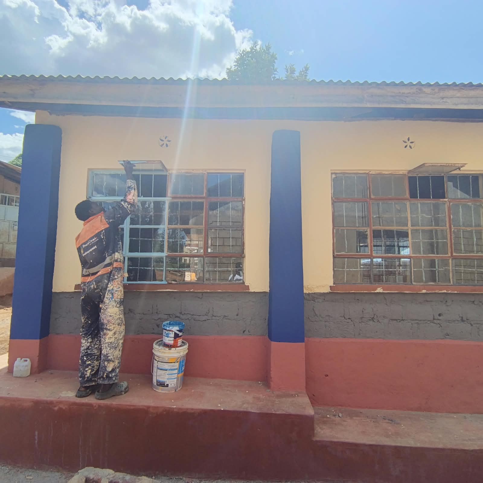 Painter refurbishing classroom window frames on a repainted block with blue pillars