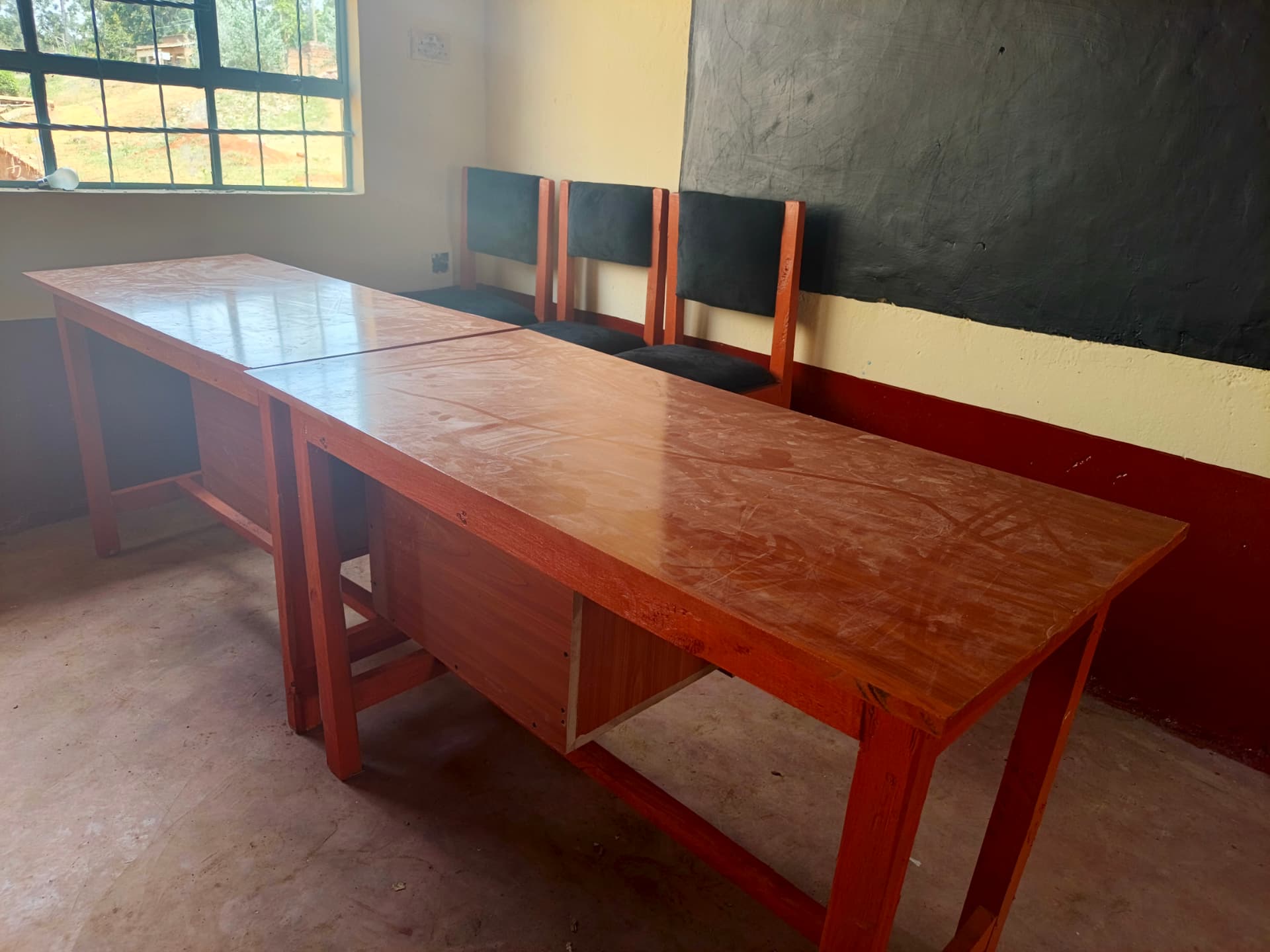 Two long teacher desks with cushioned chairs against a classroom wall